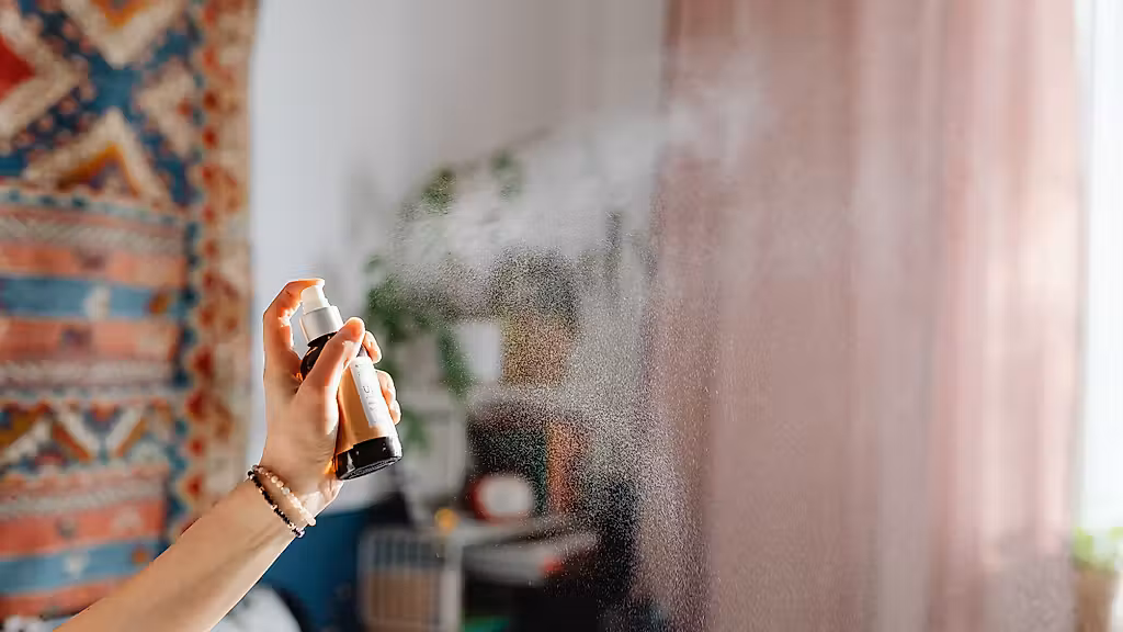 Une main pulvérisant un nettoyant en aérosol vers une fenêtre ou une surface en verre poussiéreuse, avec un léger nuage de brume et un arrière-plan de pièce flou derrière des rideaux roses.
